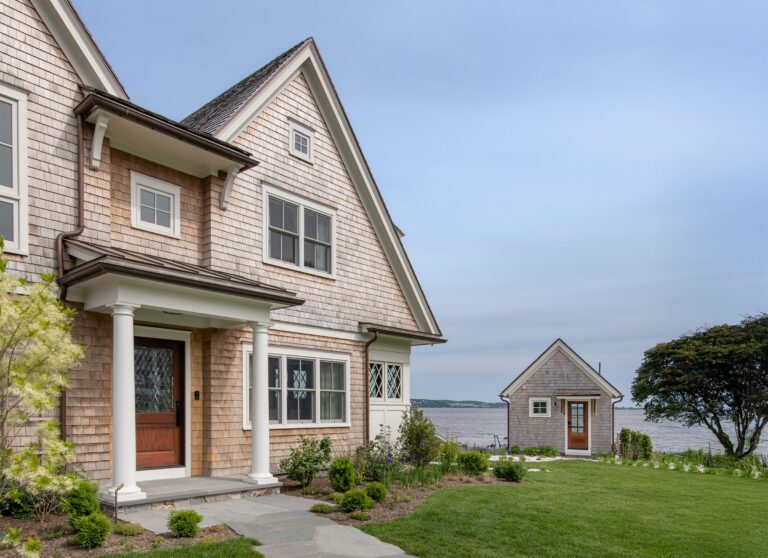 A shingle-style house with a wooden front door and white columns stands near a calm waterfront at Touisset Point. Lush green grass and shrubs surround the home, while a small matching outbuilding sits close to the water amid trees and open sky.