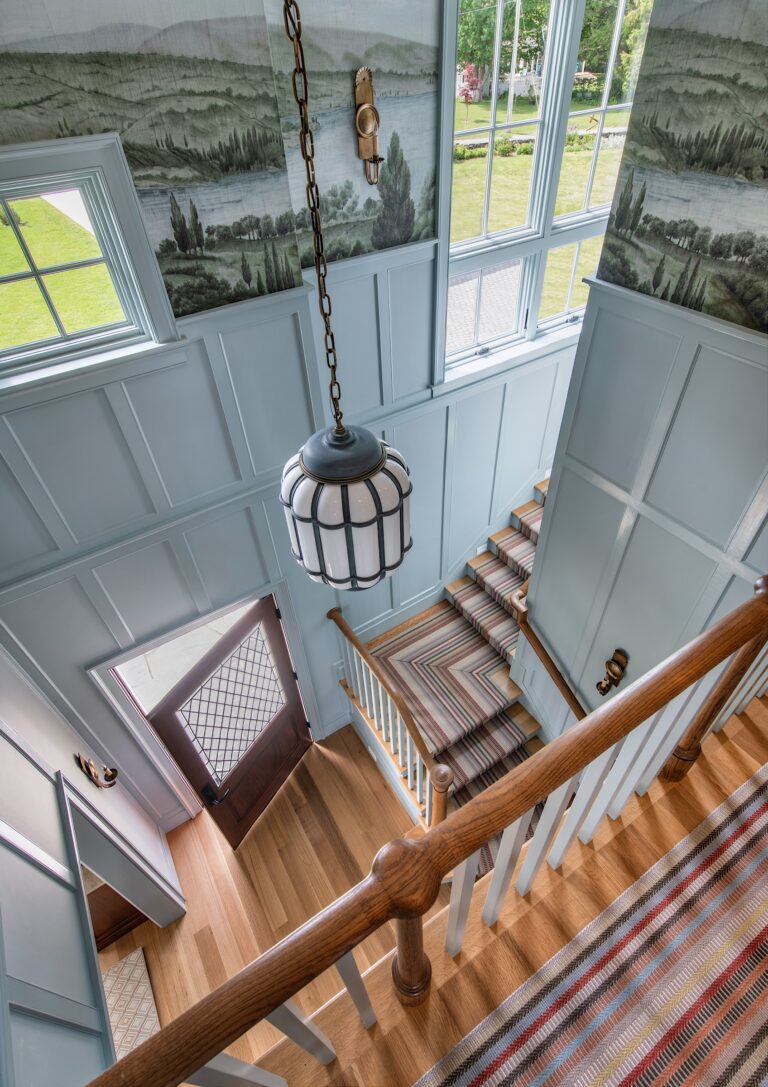A bright foyer with large windows, pale blue paneled walls, a mural of a rural landscape, and striped runner carpets on stairs and landing. A glass pendant light hangs above the open wooden front door, letting in sunlight onto the wood floor.