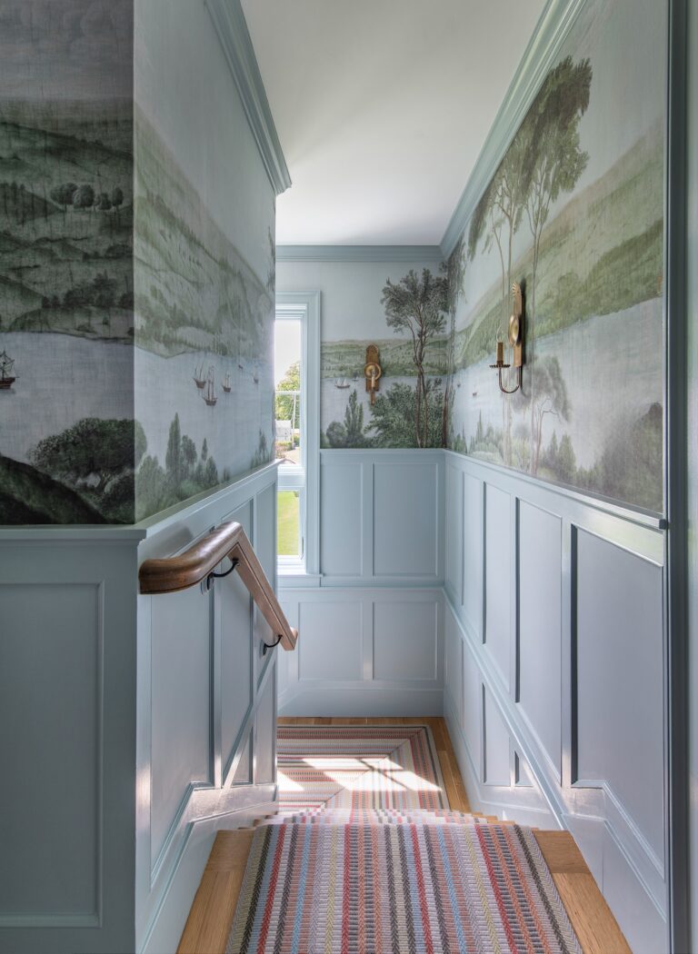 A narrow stairway landing with blue paneling, scenic wall murals of trees and water, a striped rug, a wooden handrail, and a single window letting in daylight. There is a decorative mask and brass wall sconce on the right wall.