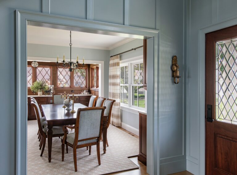 A bright, elegant dining room with a dark wood table and eight upholstered chairs, a vintage chandelier, large windows with diamond panes, striped curtains, sideboard, and decorative items. Light blue walls and wood accents create a cozy, inviting atmosphere.