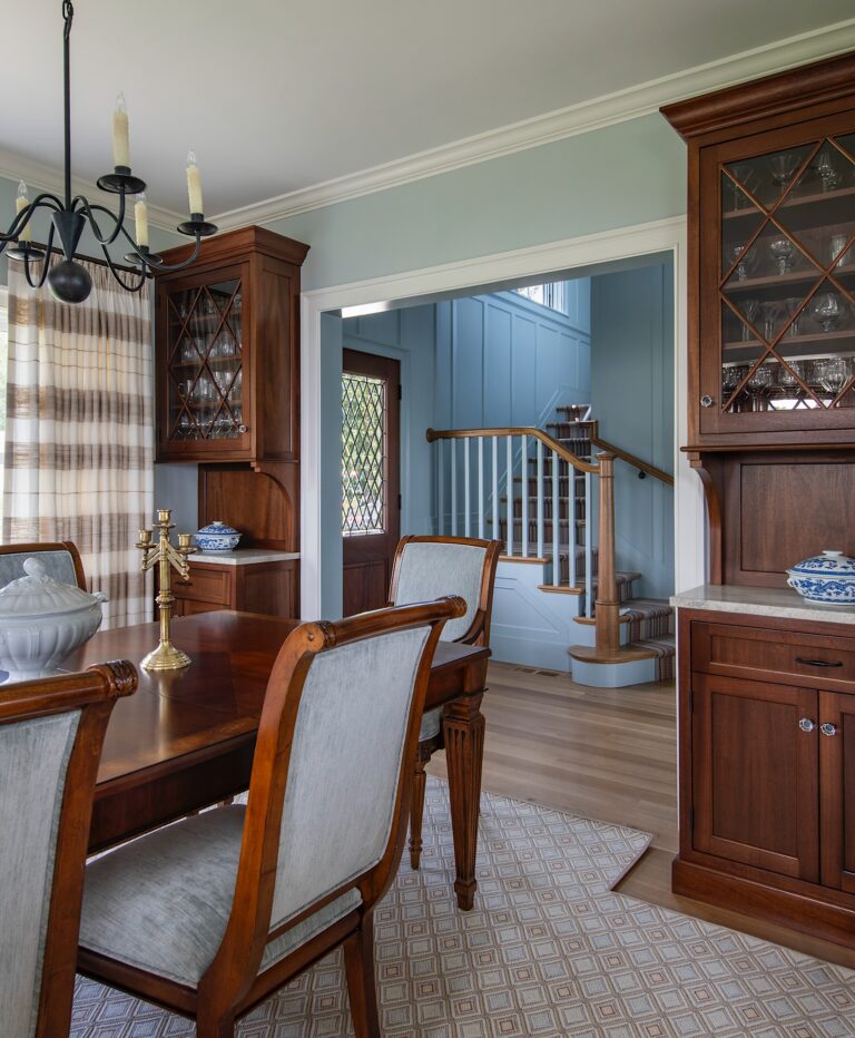 A traditional dining room with wooden table and chairs, glass-front cabinets, and a brass candelabra centerpiece. Blue and white ceramics accent the room. In the background, a staircase with wooden railing and blue paneled walls is visible. Natural light fills the space.