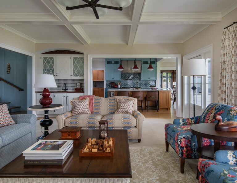 A cozy living room features patterned sofas, a blue armchair, and a wooden coffee table with books and a chess set. The room opens into a kitchen with blue cabinets, pendant lights, and barstools. Soft light enters through curtained windows.
