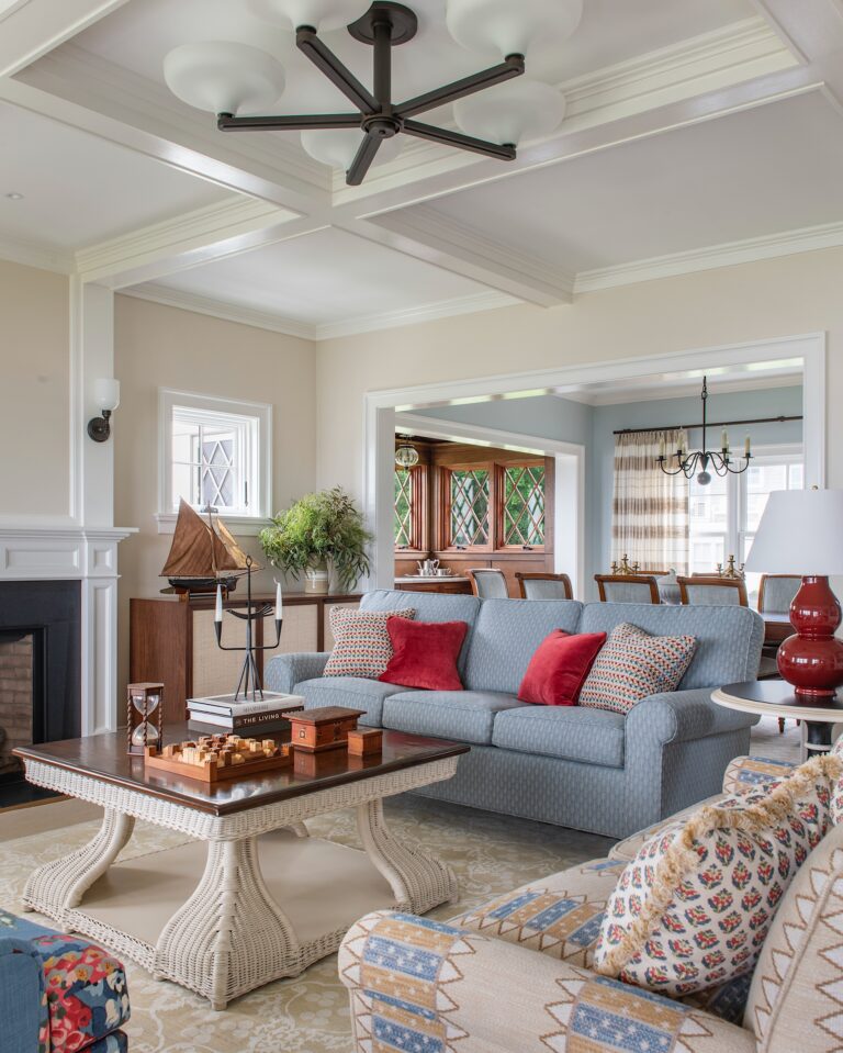 A cozy, elegant living room features a blue sofa with red and patterned pillows, a rope-style coffee table, nautical decor, and a white fireplace. In the background, a dining area with wooden furniture and a large window is visible.