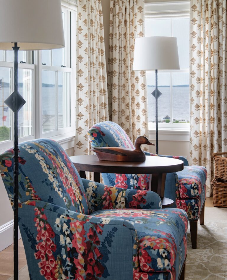 Two armchairs with blue floral upholstery face a small round table topped with a wooden duck sculpture. Floor lamps stand behind each chair. Large windows with patterned curtains overlook a body of water. A basket sits on the wooden floor to the right.