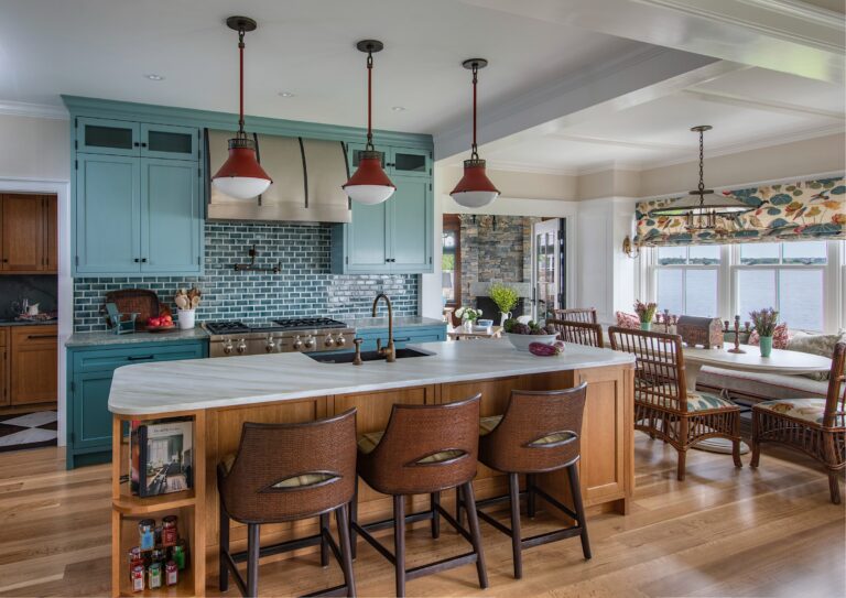 A bright kitchen features teal cabinets, a blue-tiled backsplash, stainless steel appliances, and a large island with a marble countertop and three wooden stools. Pendant lights hang above. A dining nook with wicker chairs and floral shades overlooks the water through windows.