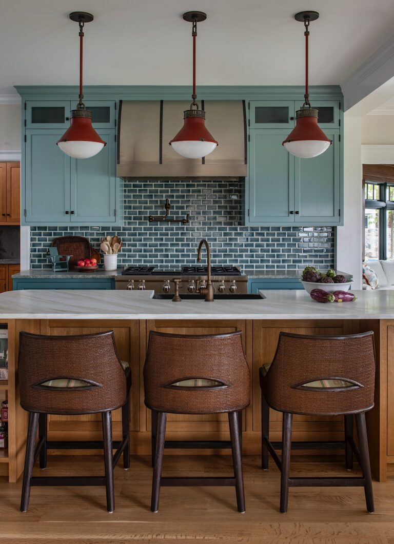A kitchen with light blue cabinets, a stainless steel range hood, blue subway tile backsplash, and a white countertop island. Three brown woven barstools sit at the island. Three red pendant lights hang above. Fruit and kitchen utensils are visible on the counters.