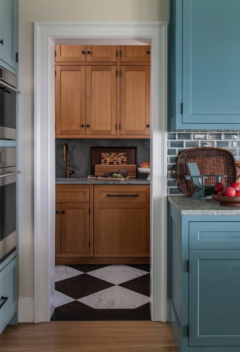 A small butler’s pantry with warm wood cabinets, a brass faucet, and dark countertop is seen through a doorway. The adjacent kitchen features blue cabinetry, a wicker basket, apples, and a gray subway tile backsplash. The pantry has a black-and-white tile floor.