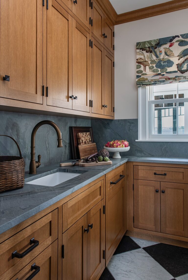 A cozy kitchen corner with honey-toned wood cabinets, dark stone countertops, and a white farmhouse sink. A bronze faucet, wicker basket, bowl of apples, and cutting board with vegetables sit on the counter. A window above features a floral-patterned Roman shade.