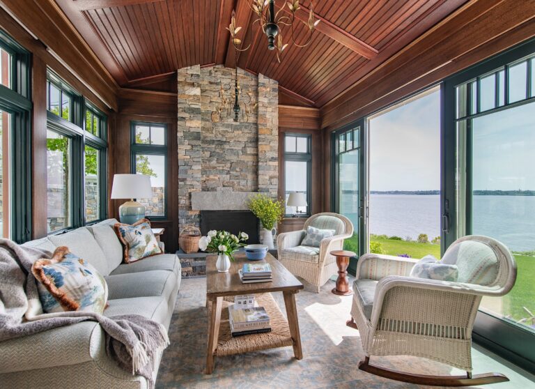 A cozy sunroom with large windows overlooking a lake, featuring wicker sofas and chairs with cushions, a stone fireplace, wooden ceiling, chandelier, coffee table with flowers and books, light rug, and serene outdoor scenery visible through glass doors.