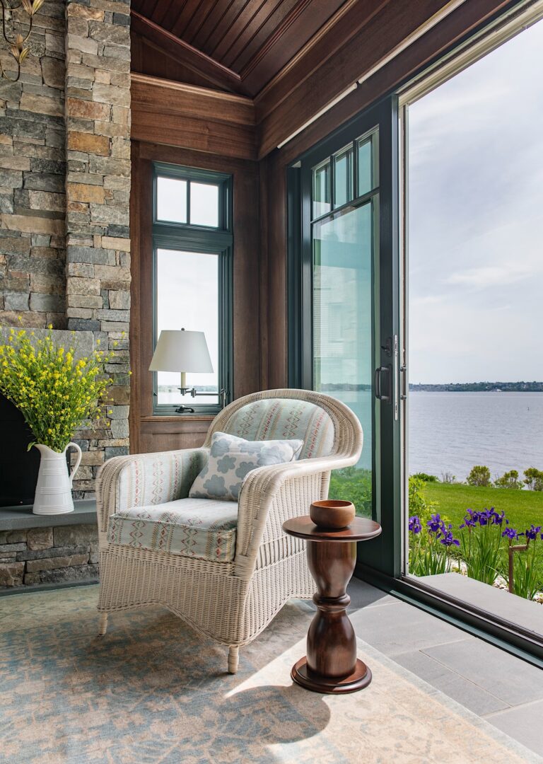 A cozy wicker armchair with a patterned cushion and pillow sits by a stone fireplace and a small wooden table, facing tall open glass doors with a scenic view of a lake, green lawn, and blooming purple flowers under a bright sky.