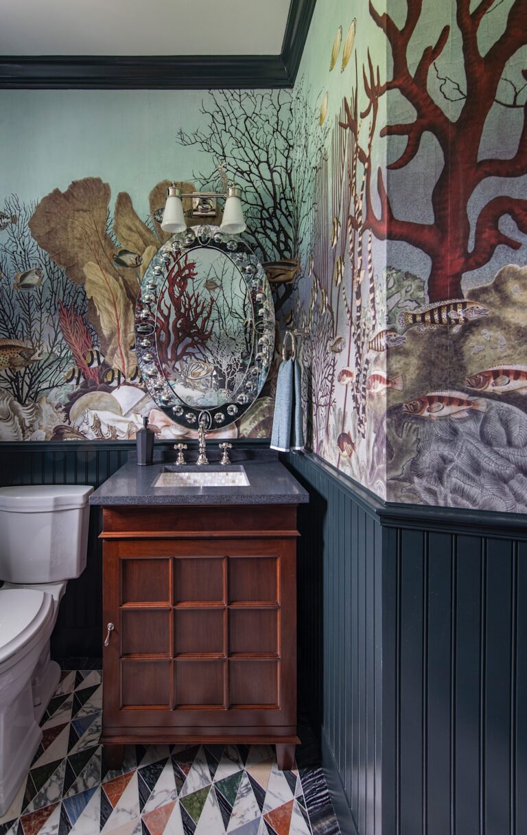 A bathroom with marine-themed wallpaper featuring coral and fish surrounds a dark wood vanity with a black countertop and oval mirror. A white toilet is beside the vanity, and the floor is patterned with multicolor geometric tiles.