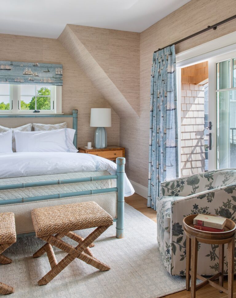 A bright, cozy bedroom with a light blue bed, white bedding, two woven stools at the foot, a wooden nightstand with a lamp, a patterned armchair, a round side table with books, nautical curtains, and a sliding glass door to a balcony.