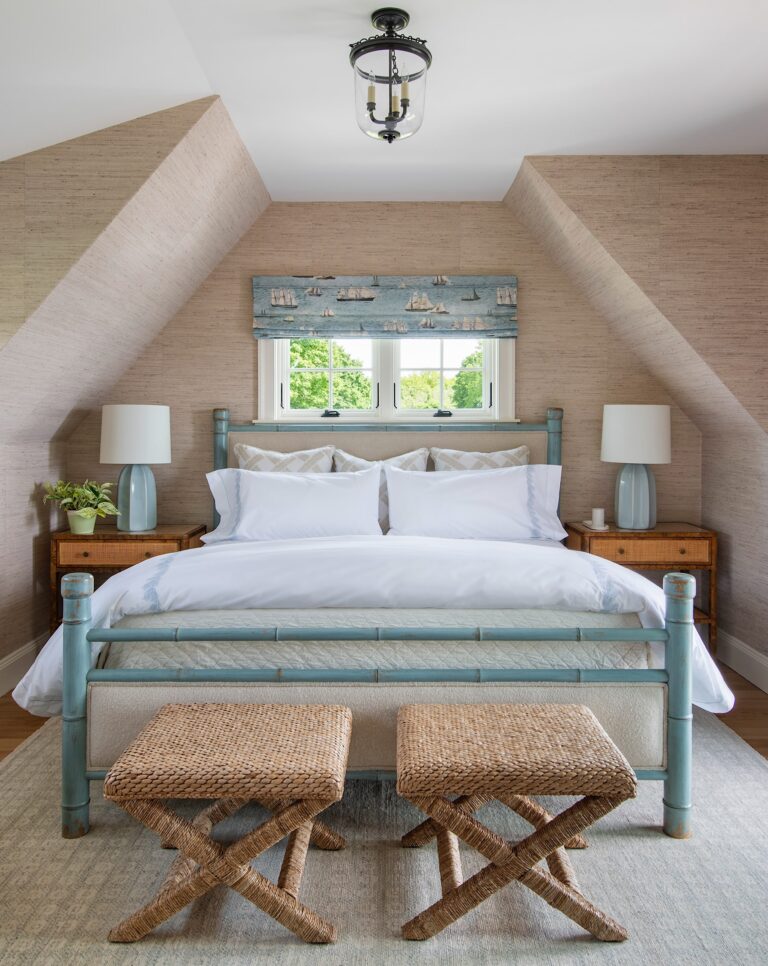 A cozy bedroom with a blue-framed bed, white bedding, and two wicker stools at the foot. There are wooden nightstands on both sides, each with a lamp and decor. A small window above the bed shows trees outside, and the sloped beige walls add to the rooms charm.
