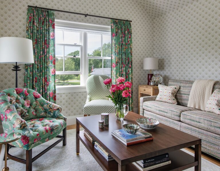 A cozy living room with floral-patterned chairs, a striped sofa with assorted pillows, a wooden coffee table holding books and pink flowers, and a lamp. Large window with floral curtains lets in daylight. Pale wallpaper and a serene outdoor view complete the scene.