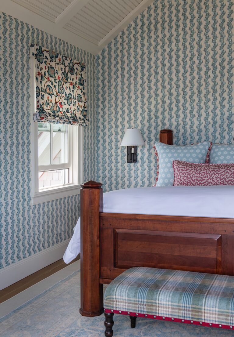 A cozy bedroom with chevron-patterned blue wallpaper, a wood bed with blue and white bedding, a floral Roman shade over the window, a plaid upholstered bench at the foot of the bed, and a wall sconce beside the bed. The room has a sloped white ceiling.