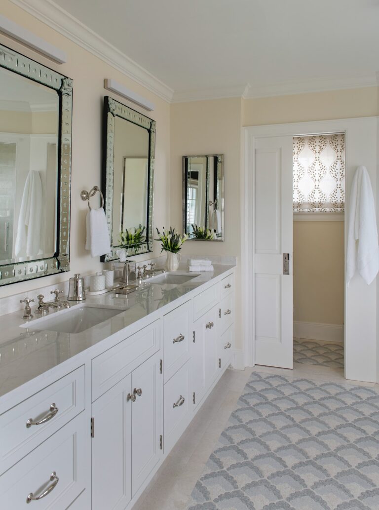 A bright bathroom with double sinks, white cabinets, two large framed mirrors, silver fixtures, and a light patterned rug. A patterned window shade filters sunlight, and small green plants decorate the countertop. Towels hang beside the mirrors and door.