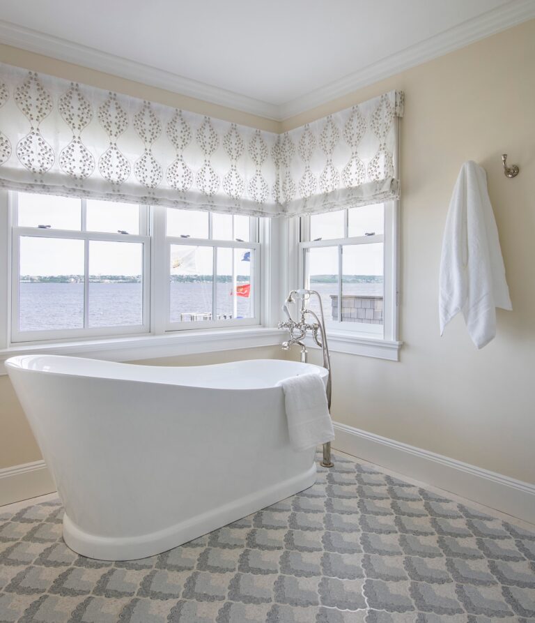 Bright bathroom with a white freestanding tub positioned by large windows overlooking water. A towel hangs on the tub and another on a wall hook. The floor has a patterned rug, and the windows have white patterned Roman shades. Neutral wall color.