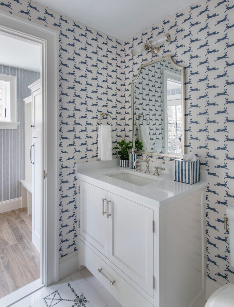 A stylish bathroom features a white vanity with a sink, chrome fixtures, and a geometric mirror. The walls have blue and white wallpaper with a leaping dog pattern. A plant and striped tissue box sit on the counter. Light wood flooring is visible in an adjoining room.